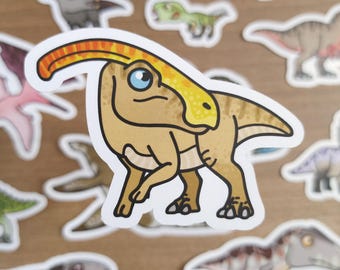 May include: A collection of dinosaur stickers. The stickers feature various cartoon dinosaurs in different colors and poses. The central sticker is a tan Parasaurolophus with a yellow crest and blue eye. Other dinosaurs include a green baby dinosaur and a brown T-Rex.