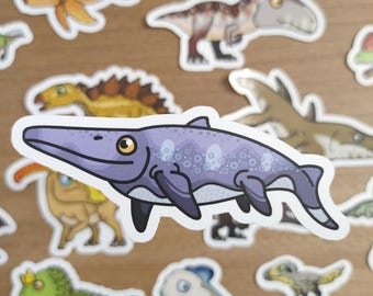 May include: A collection of cartoon dinosaur stickers in various colors. The stickers include a purple mosasaur, a green dinosaur with a crown, and other prehistoric creatures. The stickers are on a light brown surface.