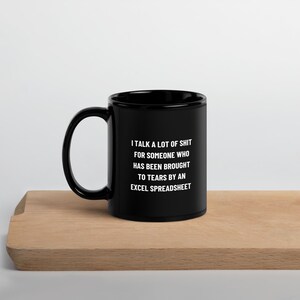 May include: Black ceramic mug with a white humorous quote: "I TALK A LOT OF SHIT FOR SOMEONE WHO HAS BEEN BROUGHT TO TEARS BY AN EXCEL SPREADSHEET." The mug is on a wooden surface.