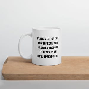 May include: White ceramic mug with a black text that reads: "I TALK A LOT OF SHIT FOR SOMEONE WHO HAS BEEN BROUGHT TO TEARS BY AN EXCEL SPREADSHEET." The mug is on a wooden surface.