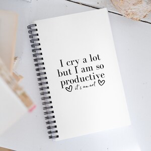 May include: White spiral bound notebook with the text "I cry a lot but I am so productive ♥ it's an art ♥" printed on the cover.