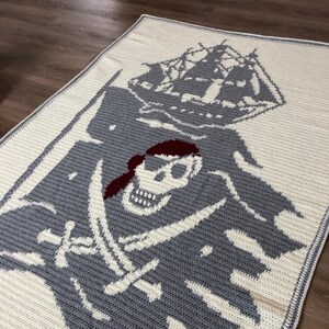 May include: A crocheted blanket with a gray and white design of a pirate skull and crossbones with a red bandana. The skull is holding two swords and a ship is in the background.