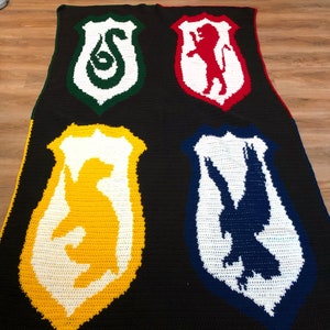 Könnte beinhalten: Häkeldecke mit den vier Wappen der Hogwarts-Häuser: Gryffindor, Hufflepuff, Slytherin und Ravenclaw. Jedes Wappen ist in einer anderen Farbe gehalten und zeigt das Haustier.