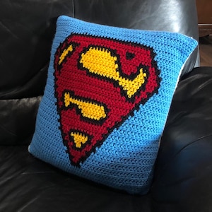 May include: A blue crocheted pillow with a red, yellow, and black Superman logo.