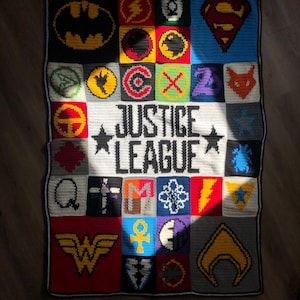 May include: A handmade crochet blanket featuring various Justice League logos and symbols. The blanket has a black border and a white central panel with the words "Justice League" in bold letters. The logos are in various colors, including yellow, red, and blue.