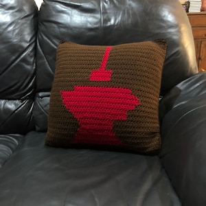 Könnte beinhalten: Ein quadratisches, gehäkeltes Kissen mit braunem Hintergrund und einem roten grafischen Design. Das Design scheint eine stilisierte Darstellung eines Gebäudes oder einer Struktur zu sein. Das Kissen liegt auf einem schwarzen Ledersofa.