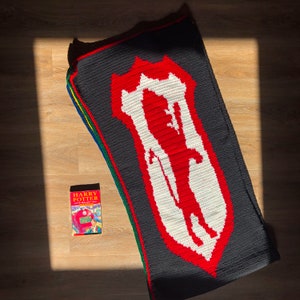 May include: A black crochet blanket with a red and white crest featuring a silhouette of a person. The blanket is folded and lying on a wooden floor. A book titled "Harry Potter and the Philosopher's Stone" is lying on the floor in front of the blanket.