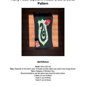 Op de afbeelding: Een haakpatroon voor het Slytherin-huiswapen uit de Harry Potter-serie. Het patroon is gemaakt met groen, wit en zwart garen. Het patroon wordt getoond op een houten oppervlak met een Harry Potter-boek.