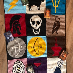 Op de afbeelding: Een gehaakt deken met vierkanten met symbolen uit de Percy Jackson-serie. Elk vierkant heeft een ander symbool, waaronder een bliksem, een schedel, een drietand en een witte roos. De boeken uit de serie liggen op het deken.