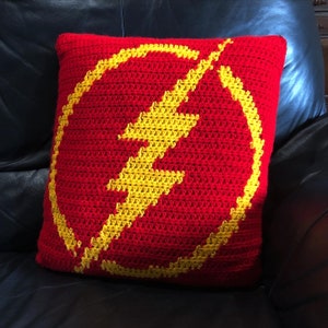 May include: A red crocheted pillow with a yellow lightning bolt design. The pillow is on a black leather couch.