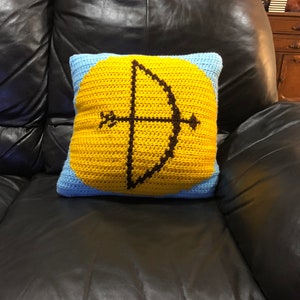 Pode incluir: Uma almofada de crochê com fundo amarelo e borda azul. A almofada apresenta um design de arco e flecha de crochê marrom e preto.