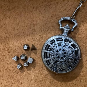 以下が含まれることがあります： 歯車が見える銀色の懐中時計と装飾的なチェーン、多面体サイコロのセットが並んでいます。サイコロには白い数字が書かれています。背景は質感のある茶色の表面です。