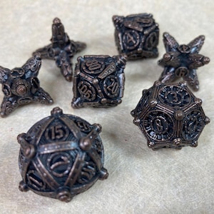 May include: A collection of bronze-colored, multi-sided dice. The set includes various shapes, such as cubes, star-shaped, and a dodecahedron. Each die features intricate designs and numbers, suitable for tabletop gaming or collecting.