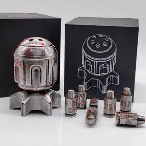 Può includere: Un oggetto metallico argentato e macchiato di rosso con una parte superiore a cupola e una base a forma di stella. Diversi piccoli oggetti cilindrici con numeri sono sparsi intorno al pezzo principale. Due scatole nere sullo sfondo.