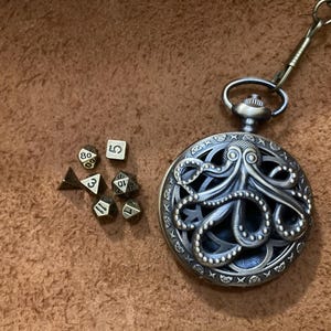 May include: A silver-colored pocket watch with an octopus design, next to a set of polyhedral dice. The watch has an intricate, openwork design. The dice are in various shapes and sizes, with white numbers.