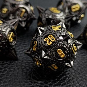 May include: Close-up of several black and gold dice with sharp, metallic spikes. The dice have gold numbers and ornate detailing. The dice are likely used for tabletop role-playing games or other dice-based games.