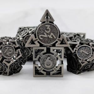 May include: A set of metallic dice with intricate geometric designs. The dice are a dark gray color and feature numbers and symbols. The dice are designed for role-playing games and tabletop games.