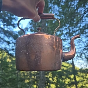 May include: A copper kettle with a brass handle and spout. The kettle is slightly tarnished and has a rustic look.