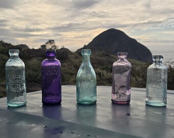 May include: Five vintage glass bottles in different colors, including blue, purple, and pink, with various labels and text. The bottles are arranged in a row on a table.