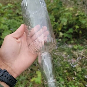 May include: A clear glass bottle with a textured surface and a long, narrow neck. The bottle is empty and has a slightly cloudy appearance.