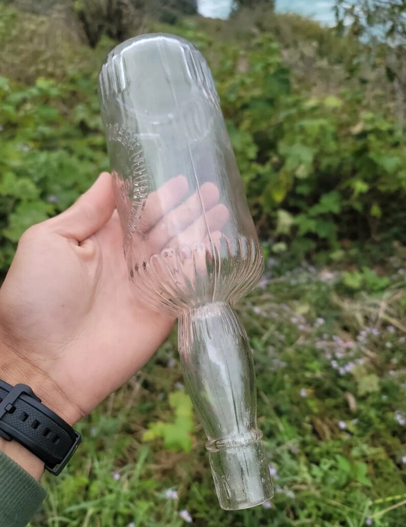 May include: A clear glass bottle with a textured surface and a long, narrow neck. The bottle is empty and has a slightly cloudy appearance.