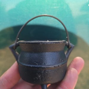 May include: A small, black cast iron cauldron with a handle and three legs. The cauldron has a rounded body and is held in a hand against a blurred green background. The handle is made of a thin, curved metal.