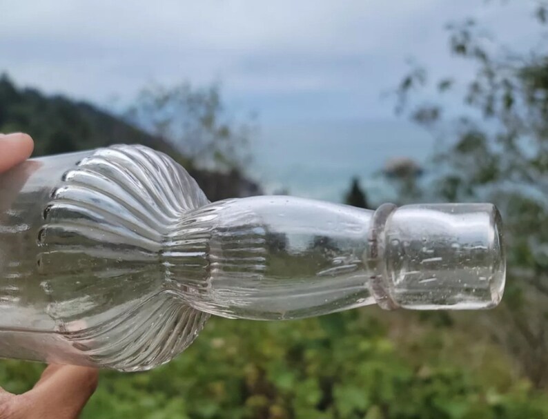 May include: A clear glass bottle with a textured, fluted design. The bottle has a round, bulbous base and a long, thin neck.