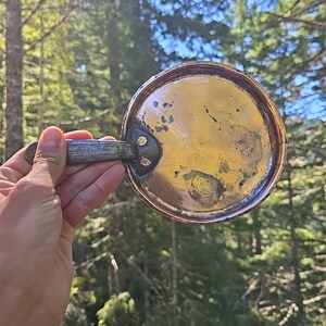 May include: A copper pan with a wooden handle. The pan has a worn, aged look and is slightly dented. The pan is held in a hand against a background of green trees.