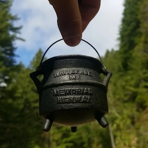 May include: A black cast iron pot with the words "Whiteface MI Memorial Highway" inscribed on the side. The pot is being held by a hand against a background of green trees.
