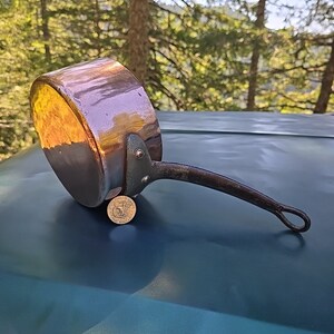 May include: A copper saucepan with a long black handle and a brass tag. The saucepan is sitting on a green surface.
