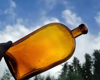 May include: A vintage amber glass bottle with a rounded rectangular shape and a narrow neck. The bottle is empty and has a slightly cloudy appearance.