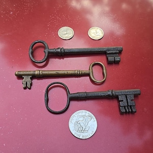 May include: Three antique keys of varying sizes and designs, alongside two small coins and a US dollar coin. The keys are made of metal, with one appearing brass-colored and the others dark. The keys are arranged on a red surface.