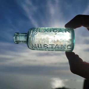 May include: A vintage, rectangular, light blue glass bottle with the embossed text "MEXICAN MUSTANG". The bottle is held up against a bright sky, with the sun shining through the glass. The bottle has a short neck and a rounded body.