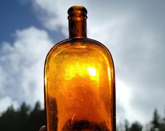 May include: A vintage amber glass bottle with a rounded body and a narrow neck. The bottle is held up against a bright sky, showcasing its translucent quality and the light passing through it. The bottle's color is a rich, warm amber.