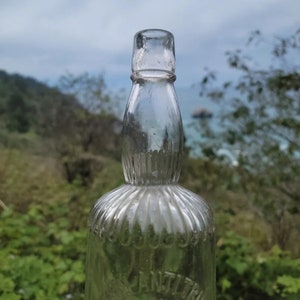 May include: A clear glass bottle with a textured surface and the word "ANTITRUST" printed on the bottom. The bottle is empty and has a vintage look.