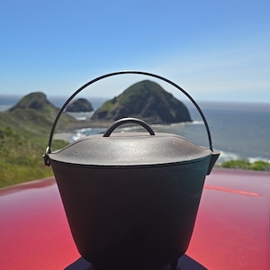 May include: A black cast iron Dutch oven with a lid and a handle. The pot has a rounded shape and sits on three small feet. A metal handle arches over the top. The background features a scenic view of the ocean and green hills under a blue sky.
