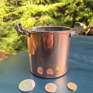 May include: A small copper pot with two handles. The pot is sitting on a blue surface with three coins next to it.