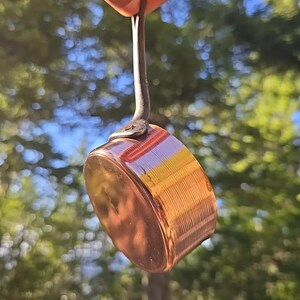 May include: A small copper pot with a long, black handle. The pot has a shiny, copper finish and is hanging from a hand.