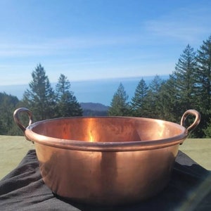 May include: A large, copper cooking pot with two handles. The pot is sitting on a black surface with a view of a blue sky and trees in the background.