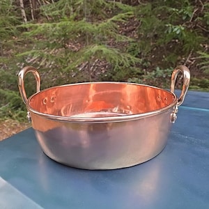 May include: A copper bowl with two handles. The bowl is shiny and has a slight patina. It is sitting on a blue surface.