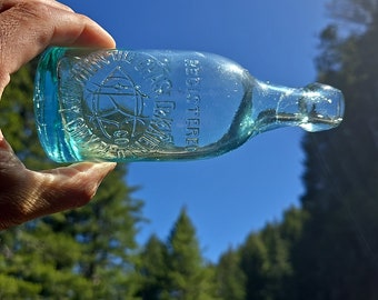 May include: A clear glass bottle with a green tint. The bottle has a rounded bottom and a long neck with a lip. The bottle is embossed with the text "Registered Trade Mark" and a star.