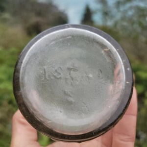 May include: Close-up of the bottom of a clear glass bottle with a dark brown lip. The bottom of the bottle has a circular embossed mark that reads "115702".