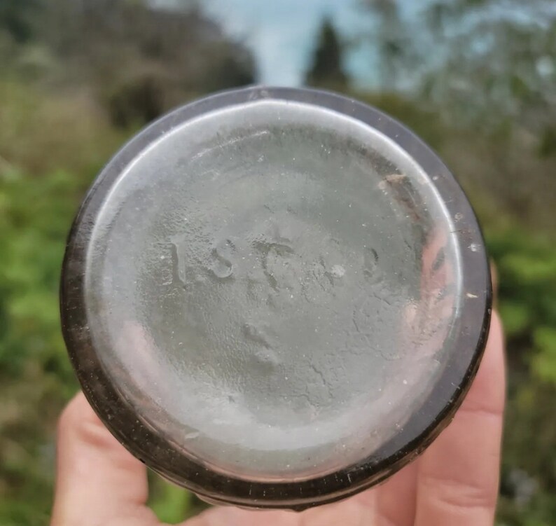 May include: Close-up of the bottom of a clear glass bottle with a dark brown lip. The bottom of the bottle has a circular embossed mark that reads "115702".