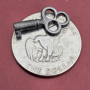May include: A small, antique-style key rests on top of a silver one-dollar coin. The coin features an eagle design and the words "ONE DOLLAR". The key is dark gray and has a decorative handle.