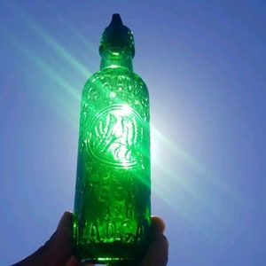 May include: A green glass bottle with a long neck and a rounded body. The bottle has embossed text and a decorative emblem. The bottle is held against a bright blue sky, with sunlight shining through it.