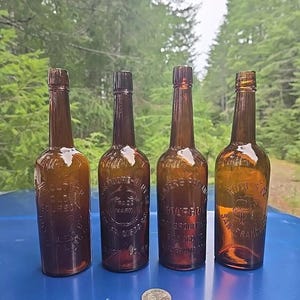 May include: Four antique glass bottles with embossed labels. The bottles are brown glass and have a variety of labels with text including "Old Bourbon", "Trade Mark", "San Francisco", and "Whiskey Straight",  "Bottled in Bond".