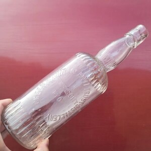 May include: A clear glass bottle with a textured surface. The bottle is embossed with the text "Green Mountain Distillery, Troy, NY, Solely Trust Anti-Colic"