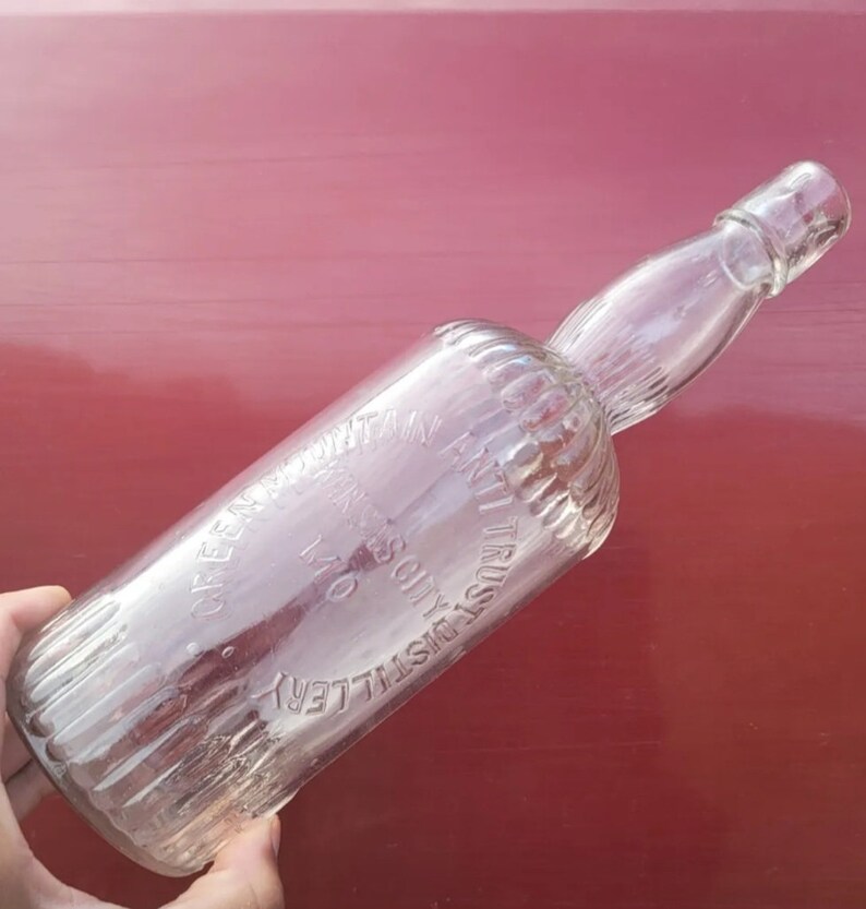 May include: A clear glass bottle with a textured surface. The bottle is embossed with the text "Green Mountain Distillery, Troy, NY, Solely Trust Anti-Colic"
