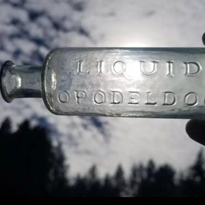 May include: A clear glass bottle with a textured surface. The bottle is labeled "LIQUID OPODELDOC".