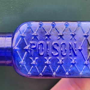 May include: A cobalt blue antique poison bottle. The rectangular bottle has a diamond pattern and the word "POISON" embossed on the front. The bottle is held between two fingers.